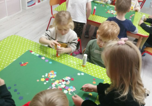Dzieci siedzą przy stolikach pokrytych zieloną ceratką i wykonują pracę plastyczną: wyklejają kwiatkami dywan dla Świętego Mikołaja.