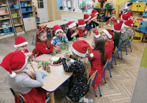 Dzieci z grupy "Słoneczka" siedzą przy stoliku, jedzą ciasteczka i piją soczki czekając na przyjście Mikołaja.