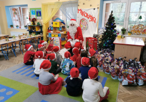 Dzieci siedzą na dywanie i słuchają o czym opowiada Mikołaj, który siedzi przed nimi.
