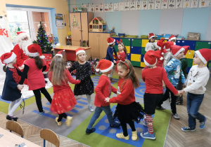 Dzieci tańczą na dywanie do słów piosenki, Mikołaj siedzi i uważnie obserwuje występ.