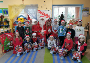 Dzieci z grupy "Słoneczek" na grupowym zdjęciu z Mikołajem i prezentami na tle dekoracji.