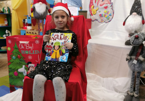 Natalia siedzi na krześle wyłożonym czerwonym materiałem i prezentuje książkę i drewnianą bombkę.