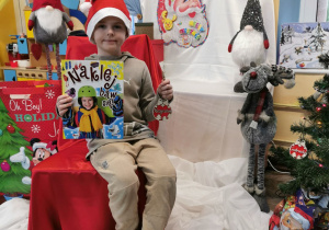 Uśmiechnięty Krzyś siedzi na tle dekoracji trzymając w ręku książkę i bombkę od Świętego Mikołaja.