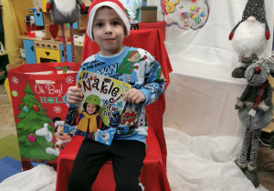Hubert K. siedzi na krześle i prezentuje książkę i bombkę od Świętego Mikołaja.
