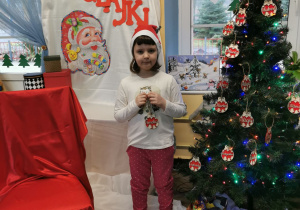 Amelia stoi na tle dekoracji i prezentuje przed sobą imienną drewnianą bombkę, którą podarował jej Mikołaj.