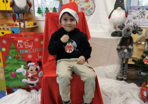Karol siedzi na krześle i prezentuje (trzymając w rączce) imienną bombkę, którą przyniósł Święty Mikołaj.