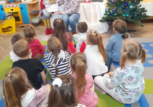 "Słoneczka" siedzą na dywanie przed mamą Gabrysi. Pani pokazuje dzieciom obrazki w książce "Wigilijna podróż Mikołaja". W tle dekoracja świąteczna.