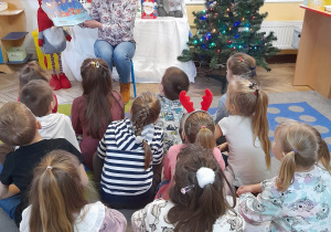 Dzieci siedzą na dywanie, a Pani czyta kolejny fragment książki pt. "Idą święta" i pokazuje obrazki. W tle dekoracja świąteczna.