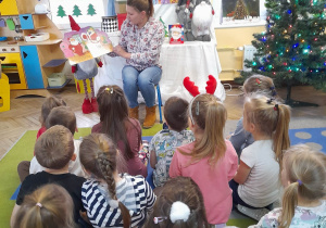 Mama Gabrysi siedzi na krzesełku na tle dekoracji świątecznej i pokazuje dzieciom ilustracje w książce pt. "Idą święta".