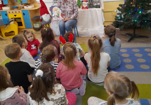 Dzieci siedzą na dywanie przed mamą Gabrysi, która pokazuje dzieciom książkę pt. "Idą święta". W tle dekoracja świąteczna - na białym materiale przypięty jest plakat z Mikołajem i reniferem. Na szafce przykrytej białym materiałem znajduje się choinka wykonana przez dzieci, świąteczny skrzat, renifer, pudełka, książka o Mikołaju. Z lewej strony na podłodze stoi świąteczny krasnal, a z prawej choinka z zapalonymi lampkami.