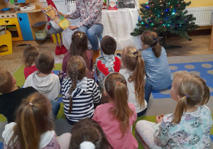 Mama Gabrysi siedzi na krzesełku, a dzieci na dywanie. Pani czyta kolejną książkę pt. "Wigilijna podróż Mikołaja".