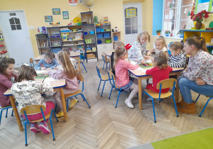 Dzieci siedzą przy dwóch stołach i rysują kredkami obrazki inspirowane wysłuchanymi opowieściami. Przy stole z prawej strony siedzi mama Gabrysi. W tle szafki z układankami, drzwi, okno.