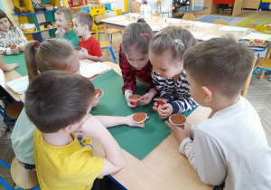 Dzieci siedzą przy złączonych stołach, na których leżą zielone kartony. Przedszkolaki oglądają stemple.