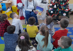 Pani z biblioteki siedzi na krzesełku, a dzieci na dywanie. Zaproszony gość prezentuje dzieciom książkę "Świąteczne opowieści" i opowiada o świętach. W tle dekoracja świąteczna.