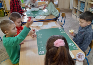 Dzieci siedzą przy złączonych stołach, na których leżą dwa kartony w kolorze zielonym oraz farby. Przedszkolaki pracują w dwóch zespołach - stemplują na kartonach tworząc wcześniej zaplanowane wzory.