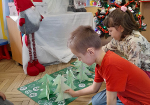 Ozdobine przez dzieci kartony leżą na podłodze obok ubranej choinki i świątecznej dekoracji. Oskar i Amelia kładą na "świątecznych dywanach" wykonane przez siebie choinki.