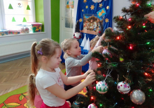 Grupa dziewczynek wiesza na choince wykonane wspólnie z rodzicami ozdoby, w tle dekoracja: na niebieskim tle szopka Bożonarodzeniowa i gwiazda Betlejemska.