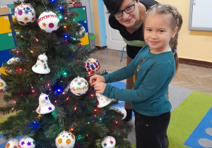 Alicja stoi ze swoją mamą obok choinki i zawiesza na bombkę na świątecznym drzewku.