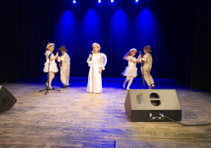 Dzieci występują na scenie. Na środku z mikrofonem w ręku stoi Alicja, a po bokach tańczą w parach Alicja i Bruno oraz Vanessa i Krzyś.