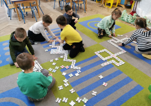 Dzieci z grupy "Słoneczek" siedzą na dywanie i układają fragment ścieżki dla ozobotów.