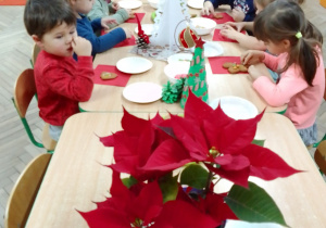 Motylki przy stolikach ozdabiają świąteczne pierniczki. Na stołach: gwiazda betlejemska, świąteczne choinki z papieru i styropianu, pierniczki, kolorowe posypki.