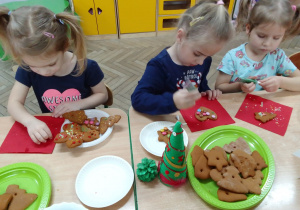 Amelia, Julia i Matylda ozdabiają pierniczki na czerwonych serwetkach, przed nimi na stoliku: świąteczne ozdoby i pierniczki.