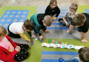Dzieci z grupy "Słoneczek" siedzą na dywanie i dopasowują kształty, które pasują do obrazków w książeczce zyg - zak.