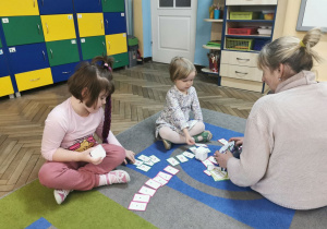 Alicja i Łucja z grupy Biedronek siedzą na dywanie, przed nimi leżą rozłożone karty do kodowania. Pani siedzi przed nimi i tłumaczy na czym będzie polegało zakodowanie GeniBotów.