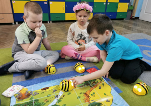 Bruno, Gabrysia i Krzysio siedzą wokół maty skarbów, która rozłożona jest na dywanie. Krzysio programuje Bee Bota tak aby cel trasy zgadzał się z poleceniem nauczyciela.