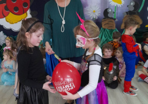 Oliwia i Alicja stoją naprzeciwko siebie. Dziewczynki trzymają czerwony balon. Obok stoi Pani Grażynka i wręcza dziewczynkom lizaki za udział w konkursie. W tle kilkoro dzieci z innych grup oraz dekoracja balowa.