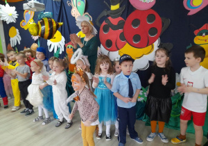 Dzieci w strojach balowych stoją razem z Panią Grażynką na tle dekoracji i klaszczą w ręce przy muzyce.