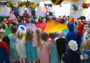 Dzieci ze wszystkich grup stoją w kole i trzymają chustę animacyjną. Obok dzieci stoi wodzirej "Alex" z mikrofonem w ręku. W tle dekoracja balowa.