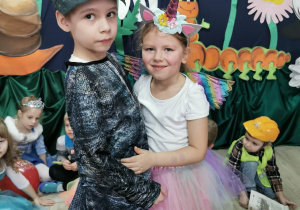 Hubert w stroju Dinozaura i Gabrysia w stroju Jednorożca stoją obok siebie podczas konkursu "Taniec na gazecie". W tle kilkoro dzieci z innych grup przykucnęło na podłodze oraz dekoracja balowa.