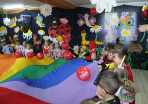 Dzieci trzymają chustę animacyjną i podrzucają czerwone balony przy muzyce. W tle dekoracja balowa.
