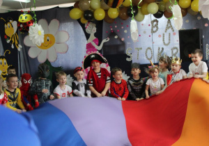 Grupka dzieci ubrana w stroje balowe stoi obok siebie trzymając chustę animacyjną. W tle dekoracja.