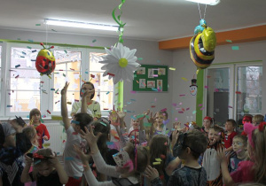 Dzieci stoją w rozsypce na środku sali, a obok Pani Dyrektor. Przedszkolaki łapią kolorowe konfetti. W tle okna, a przy suficie wiszą balony w kształcie pszczoły i biedronki oraz papierowa biała stokrotka.