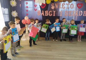 Dzieci z grupy Biedronek stoją na tle dekoracji z okazji Dnia Babci i Dziadka. W dłoniach trzymają upominki przygotowane dla dziadków.