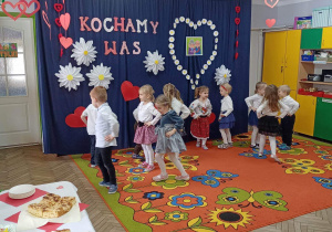 Dzieci na tle dekoracji prezentują układ taneczny do piosenki „Nazywają mnie Poleczka”. Z lewej strony stół nakryty białym obrusem, na którym są talerze z ciastem drożdżowym.