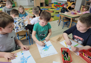 Dzieci z grupy Biedronek siedzą przy stolikach w trakcie kolorowania obrazków według podanego kodu.