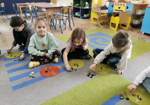 Igor, Vanessa, Winek i Oskar siedzą na dywanie, przed nimi leżą kolorowe krążki na których układają określoną liczbę guzików.