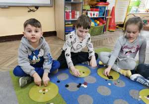Karolek, Hubert i Ala siedzą na dywanie. Przed nimi leża kolorowe krążki, na których układają określoną liczbę guzików podczas zabawy z guzikami.