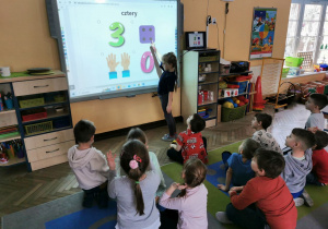 "Słoneczka" siedzą na dywanie przed tablicą multimedialną. Natalia stoi przed tablicą i rozwiązuje zadanie, wskazuje cyfrę.