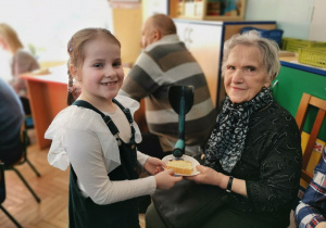 Na zdjęciu uśmiechnięta Ala podaje babci ciasto na talerzyku.