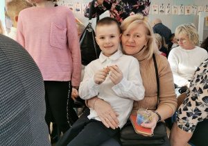 Uśmiechnięty Krzysio siedzi na kolanach u babci trzymając w ręku cukierka.