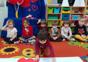 Filip układa połówki czerwonych serduszek na dywanie. Wokół siedzą Motylki i obserwują działania kolegi. Z tyłu dekoracja: na niebieskim tle napis Walentynki, czerwone serca i balony w kształcie serc.
