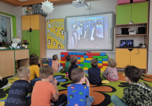 Biedronki siedzą na dywanie przed tablicą multimedialną oglądają film edukacyjny o pingwinach