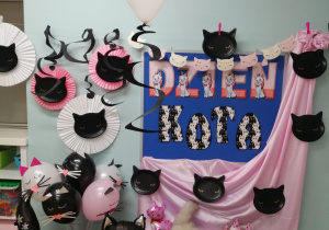 Dekoracja z okazji Dnia kota: na tablicy znajduje się napis Dzień kota,po jednej stronie tablicy wisi różowy materiał na którym przypięte są czarne głowy kotów, a po drugiej stronie stoi wazon z balonami w kształcie kotów oraz czarny balonowy kot. Na szafce pokrytej różowym materiałem leżą papierowe czapeczki kotów, kocie gwizdki oraz leży biały , puszysty kot.