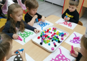 Grupka dzieci siedzi przy stoliku, na którym znajdują się na białej tacy kolorowe pomponiki. Każde dziecko ma plansze z kolorowymi kotami i spinacz do bielizny.