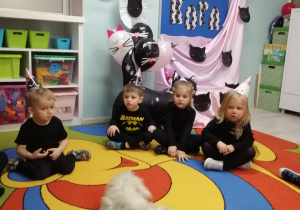 Grupka dzieci siedzi na dywanie , na środku leży biały , puszysty kot. W tle dekoracja z okazji Dnia kota.