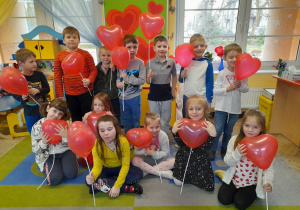 Dzieci pozują do zdjęcia trzymając czerwone balony w kształcie serca. W tle tablica, na której przypięte są sylwety serc.